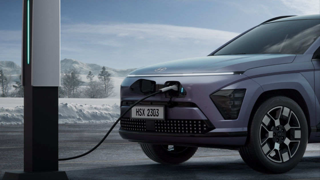 A photo of the left quarter view of The all-new KONA Electric being charged via a charging station, with a snowy background in the background.